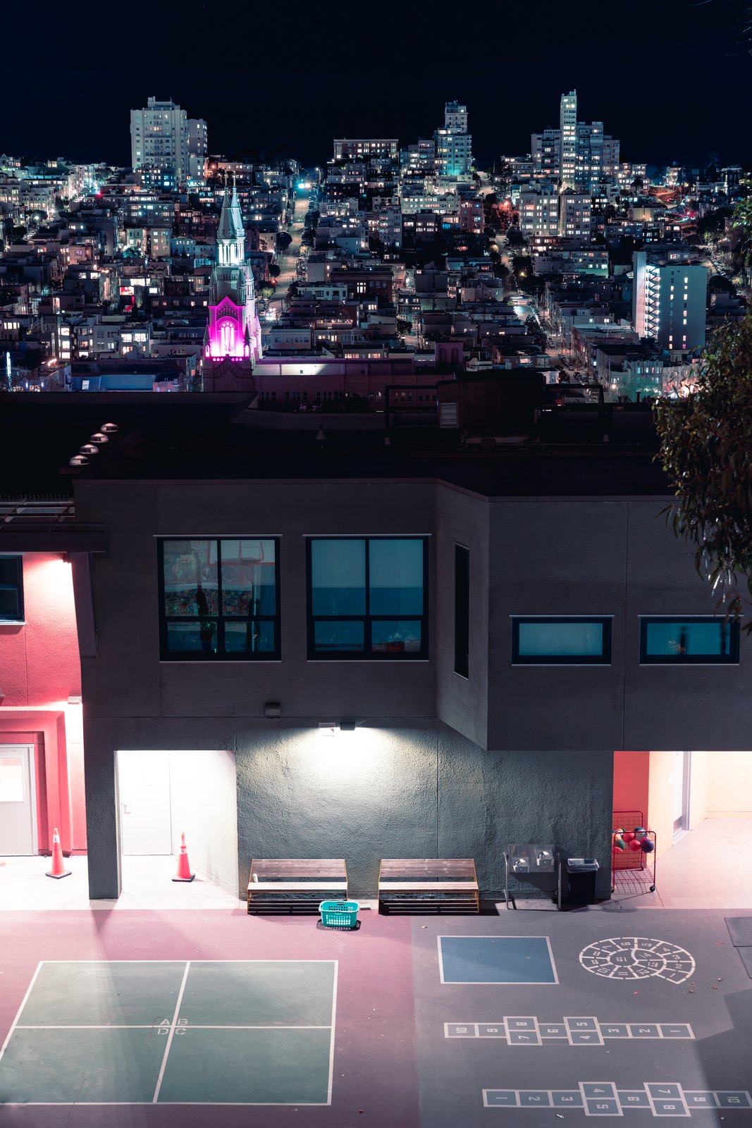 A schoolyard at night with a magenta-lit church tower and city lights behind it