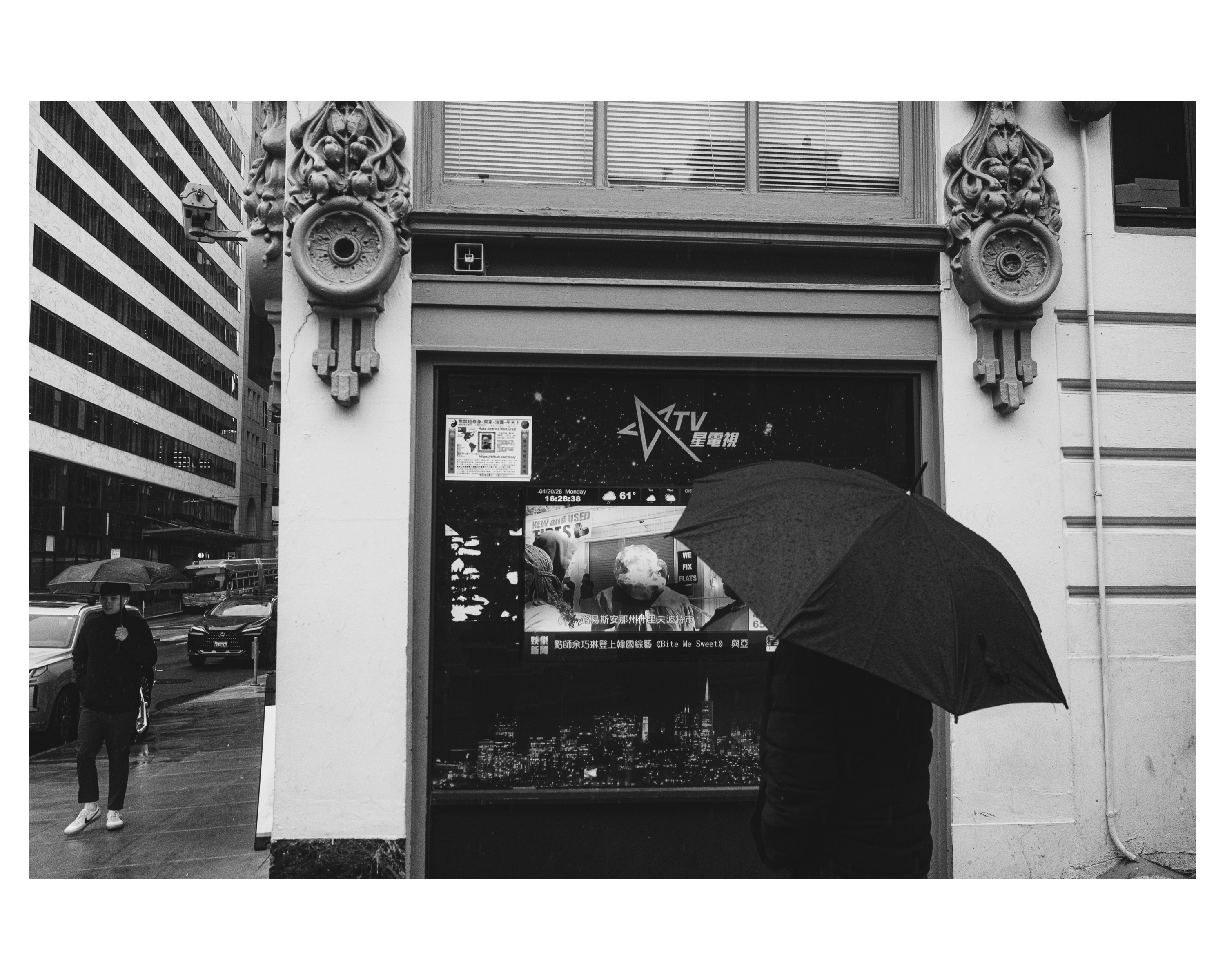 A person under an umbrella passing a storefront TV screen on a rainy day