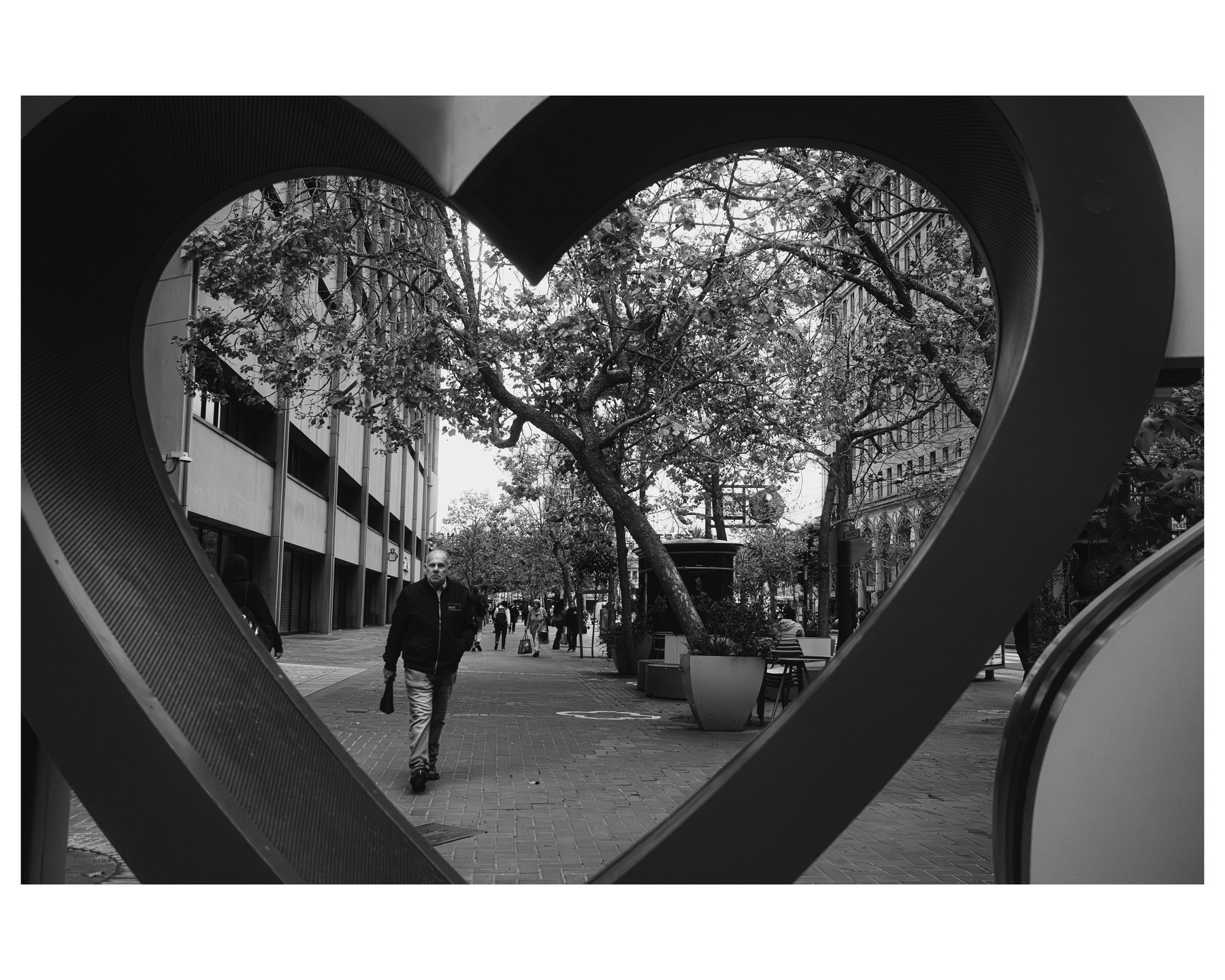 A man walking down a tree-lined street, framed through a large heart sculpture